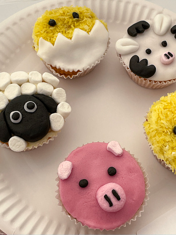 Stock photo showing close-up, elevated view of a set of farm animal themed decorated cupcakes on white, paper plate.