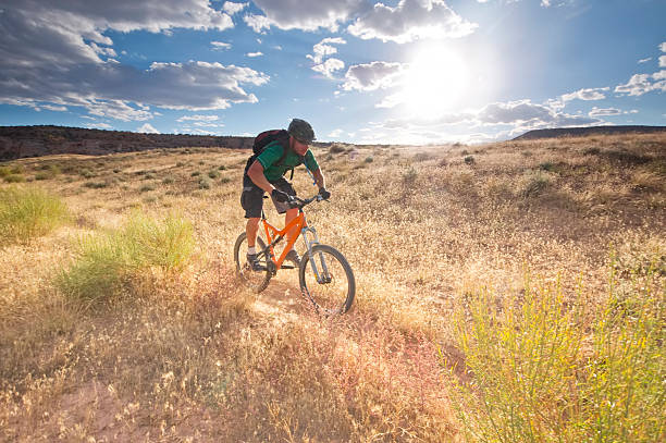 outdoor sports and adventure mountain biker speeding through the desert meadow terrain beneath a cloud filled blue sky filled with the sun during sunset turning the landscape a golden yellow. horizontal composition with copy space.  fruita, colorado. fruita colorado stock pictures, royalty-free photos & images