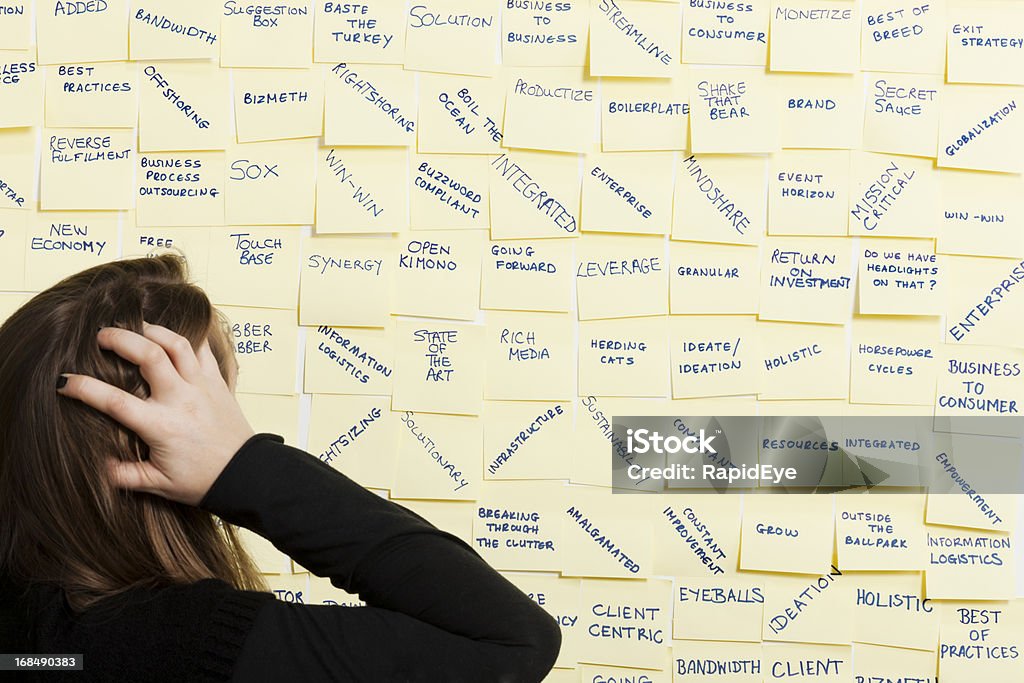Worried woman looks at board full of business buzz words Back view of confused woman scratching her head as she looks at a noticeboard covered with business jargon and buzz words witten on yellow adhesive notes. Technophobe Stock Photo