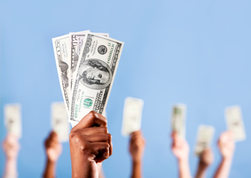 A group of hands of mixed sexes and races, all holding up US dollars. Focus is on the African man's hands in the foreground. 