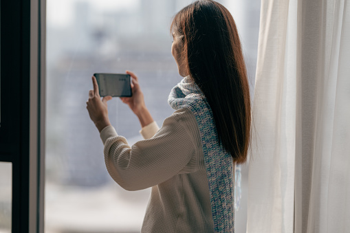 Young beautiful Asian woman standing near the window with neck scarf and using smart phone indoor.
