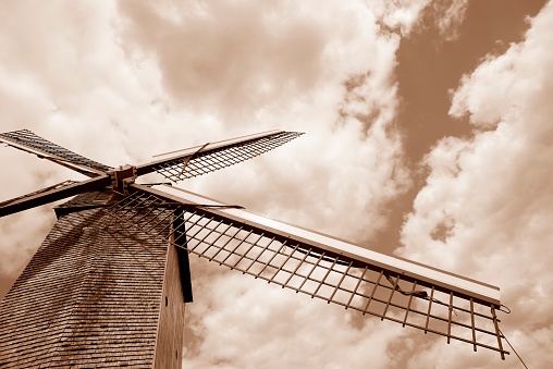 A sepia toned image of a traditional windmill in Bruges, Belgium, on a bright day with voluminous clouds passing by.