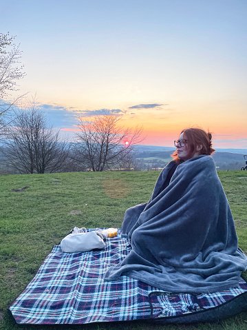 Stock photo showing close-up view of an attractive, red haired young woman sitting on a picnic blanket wrapped in a blanket as part of an off-season, sunset picnic, romantic date.