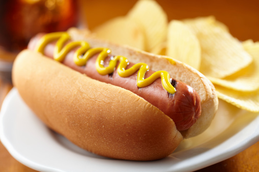 Hot dog with mustard and ripple chips on plate with cola in background, shot with very shallow focus.