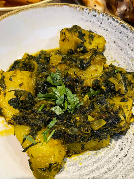 Stock photo showing an elevated view of a restaurant meal of Saag aloo, cubed potatoes and spinach flavoured with spices, cumin, garlic, green chillies, onion, turmeric, ground coriander and fresh root ginger. imagen en primer plano del entorno del restaurante de un plato que contiene saag aloo (papas en cubos con verduras), espinacas aromatizadas con especias, comino, ajo, chiles verdes, cebolla, cúrcuma, cilantro molido y jengibre de raíz fresco, fondo de me