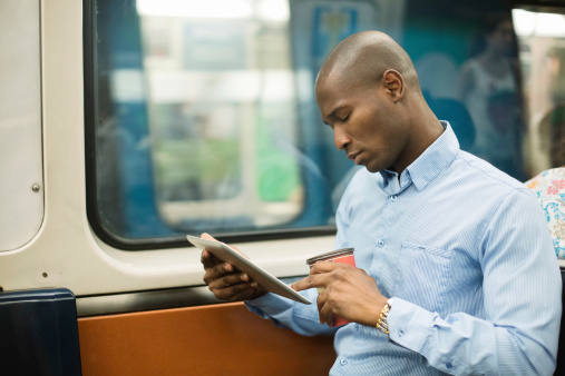 African man  commuting on subway while working with digital tablet. He has a coffee in his other hand.