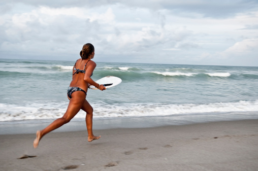 A 19-year-old woman running to catch a wave with her skimboard. Some motion blur. Photographed at sunset/dusk.