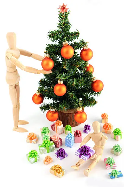 family scene of two wooden mannequin. The big one is decorating the with christmas tree. The little one sits under the tree looking exciting at the christmas presents. 