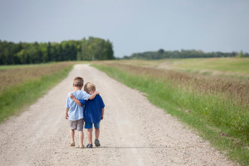Two boys hugging and walking down a rural gravel road. Rear view. Caucasian models. Young children are four and five years old. Themes include friendship, love, care, brothers, siblings, friends, bonding, fun, walking, consoling, cute, relationships, mates, talking, comfort, and summer. 
