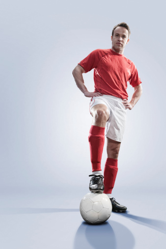 A studio portrait of a soccer player standing with hands on hips and one foot on a football. The player is dressed in a generic red and white soccer kit. Shot in the studio against a plain white background with copyspace.
