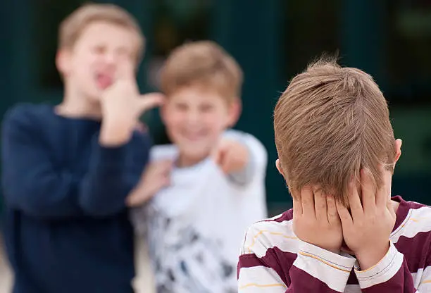 An upset elementary school boy hides his face while being bullied by two other boys.  Shot in front of their elementary school.