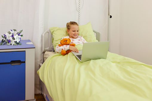 Young Patient Girl Sitting in Pediatric Hospital Bed While Watching Funny Cartoons. Sick Little Child Using Computer to Watch Comical Content on Internet While Sitting in Pediatric Clinic Ward Room.