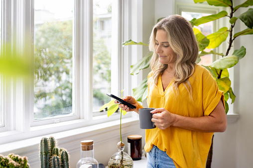 Happy woman at home looking at social media on her cell phone while drinking a cup of coffee - lifestyle concepts