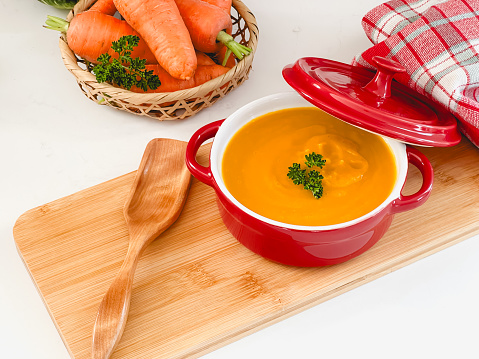 Cream of carrot soup, in red pot, on a bamboo board, on white countertop, copy space