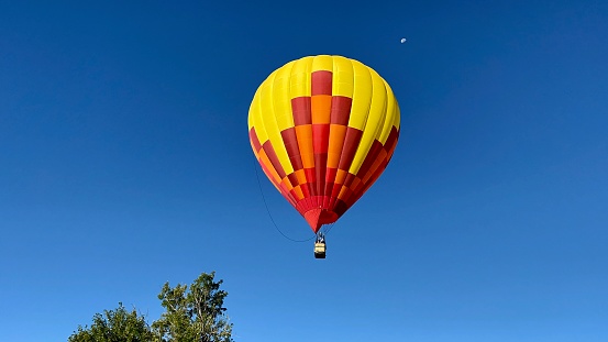 Yellow and red hot air balloon flies in air above tree tops in early morning sky with half moon visible above balloon in Colorado