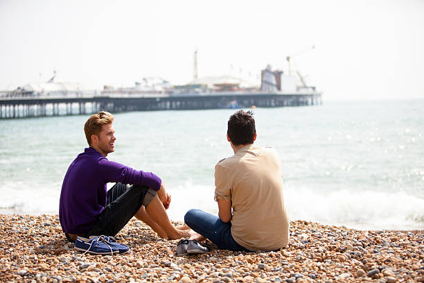 Gay male couple sitting on the beach in Brighton A handsome gay couple sitting together relaxing and chatting on the beach on a sunny day in Brighton, England. brighton england stock pictures, royalty-free photos & images