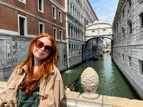 Stock photo showing close-up view of red haired woman standing on bridge over the Rio di Palazzo with Ponte dei Sospiri (Bridge of Sighs) in the background with tourists in gondolas piloted by gondoliers travelling along the canal.