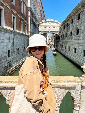Stock photo showing close-up view of red haired woman standing on bridge over the Rio di Palazzo with Ponte dei Sospiri (Bridge of Sighs) in the background with tourists in gondolas piloted by gondoliers travelling along the canal.