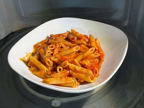 Stock photo showing close-up, elevated view of white, ceramic bowl containing penne pasta covered in rich tomato sauce on glass turntable in microwave oven.