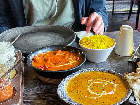 Stock photo showing close-up view of bowls of saffron pilau rice, Butter chicken and Dal makhani (black bean curry) served with a tray of triangle quarters of naan flatbread and triangle quarters of naan flatbread in a restaurant.