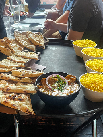 Stock photo of bowls of saffron pilau rice and rectangular trays of triangle quarters of naan flatbread on server's folding side table tray set beside restaurant table of customers.