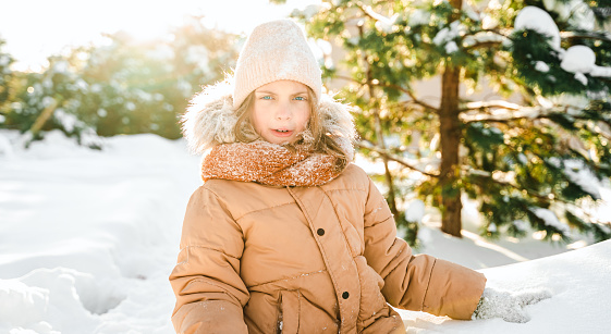 Little cute girl in beige hat,orange scarf snood is lying,playing in snow. Kid walking in beautiful frozen forest park among snowy trees. Winter vacation entertainment. Village country house backyard.