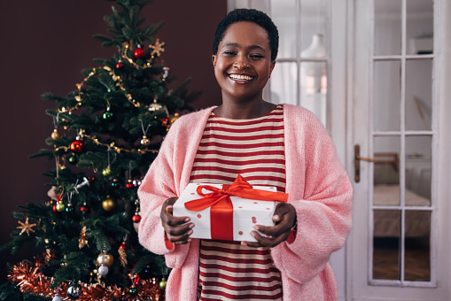 A portrait of an Afro-American woman holding a Christmas present in her living room, smiling.