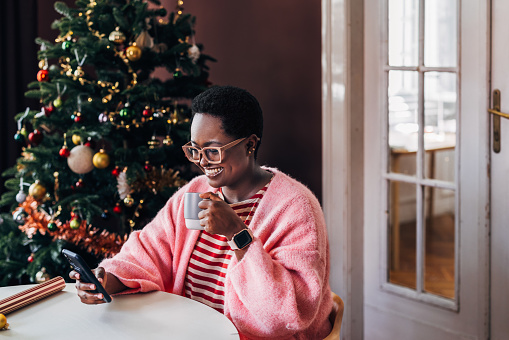 An Afro-American woman enjoying her Christmas holiday, drinking coffee and browsing her social media on a mobile phone device.