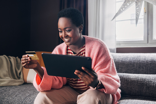 Afro-American woman sitting casually on the sofa in her living room, shopping online using her credit card and tablet device.