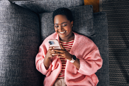 An Afro-American woman sitting comfortably on the sofa in her living room, using her cell phone device, enjoying Christmas holiday.