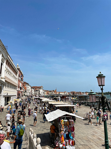 Venice, Italy - July, 12 2023: Stock photo showing close-up view of tourists milling around the water taxi area and old buildings in paved waterfront area of Venice, Italy with market stalls of outdoor market on Riva degli Schiavoni.