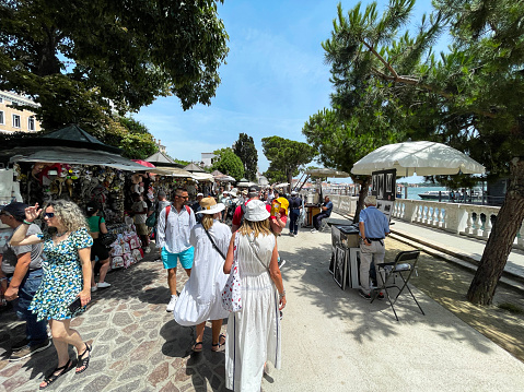 Venice, Italy - July, 12 2023: Stock photo showing close-up view of tourists milling around paved waterfront area of Venice, Italy near Giardini Reali with market stalls of outdoor market.