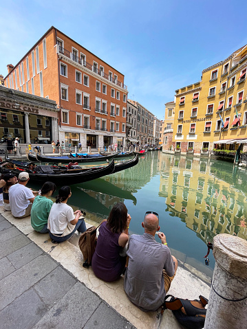 San Marco district, Venice, Italy - July 12, 2023: Stock photo showing close-up view of tourists sat on steps at the Orseolo Basin near the Hard Rock Cafe,  waiting for a gondola trip. This small lagoon was opened in 1863 to create a landing for gondolas near the heart of the city.