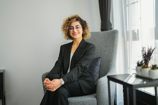 Smiling businesswoman in black jacket with curly hair sitting on sofa in office