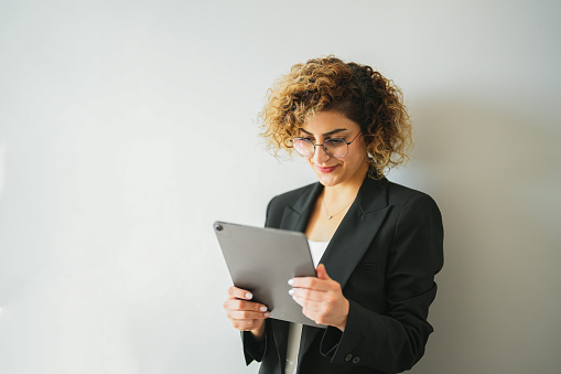 Businesswoman with curly hair and black jacket in front of white background looking at her tablet in the office