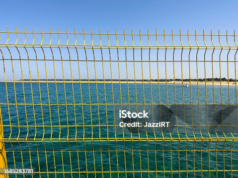 istock Blue sky behind a wire fence. Beach and sea in the background. Sunny summer day. Yellow wire mesh for security on the ferry pier. 1685283781