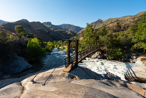 Hanging wooden bridge in Potwisha campground, in Sequoia National Park. A landscape of trees, mountains and rocks, and a fast river passes under the bridge