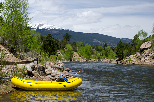 An yellow river raft sits empty on the side of the Arkansas River in Buena Vista, Colorado. After a year of heavy snows in the high country, the river runoff is high, and it appears that this group of rafters has taken time away from the whitewater to enjoy a picnic away from their boat.  The snow covered Collegiate Peaks Range stands in the background as the river meanders through the image.