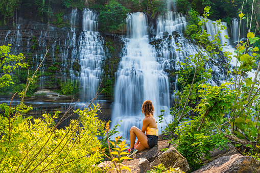 Attractive young woman gazing up at large waterfall with lush foliage in soft golden light. Shot in Tennessee, USA.