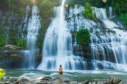 Attractive young woman gazing up at large waterfall with lush foliage in soft golden light. Shot in Tennessee, USA.