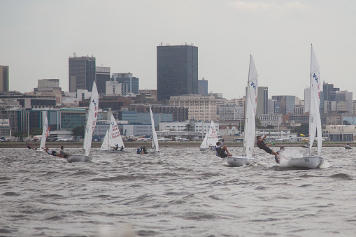 Rio de Janeiro, Brazil – September 10, 2023: several sailors in the 420 class sailboat regatta on a very windy and rainy day in Guanabara Bay during the Rio de Janeiro sailing week. In the background the city of Rio de Janeiro financial district.
