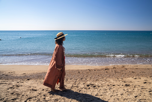 Full length photo of woman wearing a straw hat and a beachwear walking on sands. Shot under daylight with a full frame mirrorless camera.