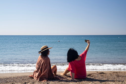 Photo of two women wearing long beachwear sitting on beach sand and taking selfie. Shot under daylight with a full frame mirrorless camera.