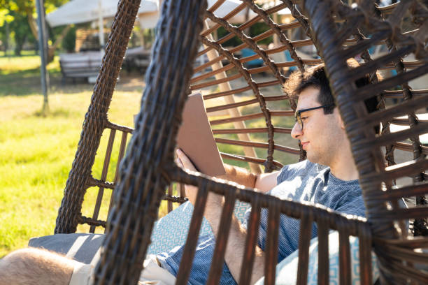 Photo of young man sitting on bamboo hammoch style chair and reading a book. Shot under daylight with a full frame mirrorless camera.