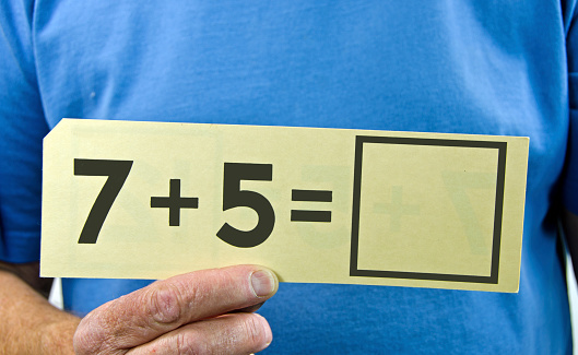 A man's hand holding an old educational  addition flash card with the numbers 7 + 5. Blue shirt background with copy space.