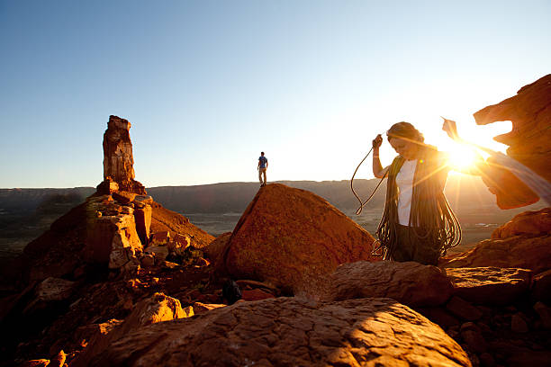 A happy young female rock climber coils her climbing rope at sunset as her husband stands on a rock in the distance looking for the trail back to camp.
