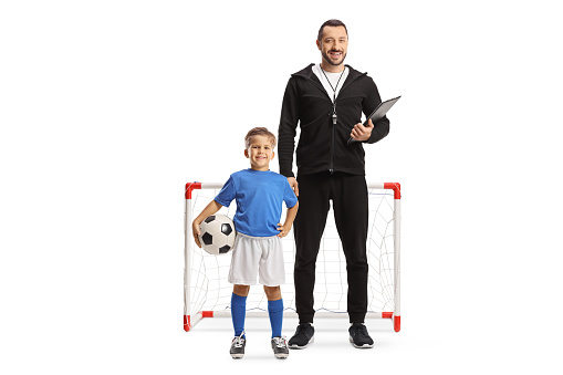 Football coach with a whistle holding a clipboard and a boy standing next to him in front of a mini goal isolated on white background