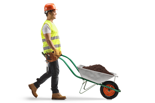 Full length profile shot of a construction worker pushing a wheelbarrow with soil isolated on white background