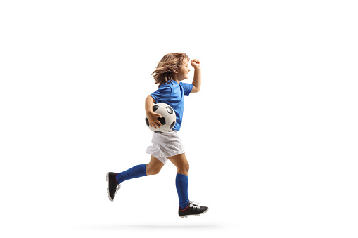 Full length profile shot of a girl in a football kit running with a ball isolated on white background