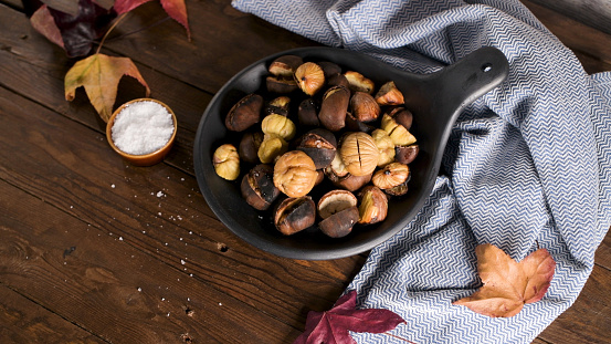 Roasted chestnuts in cast iron pan over rustic wooden board and grey wooden background, selective focus.